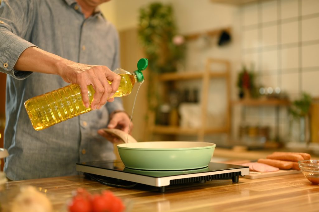 Senior man pouring cooking oil into a green frying pan while preparing to cook in warm kitchen